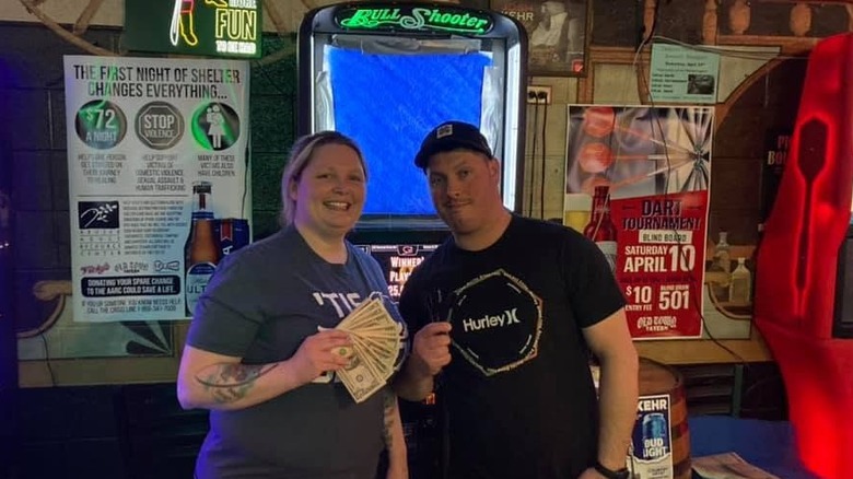 A smiling woman (left) holding a fanned out wad of cash and a man (right) holding darts stand in a bar in front of a Bull Shooter dart machine surrounded by random posters and clutter