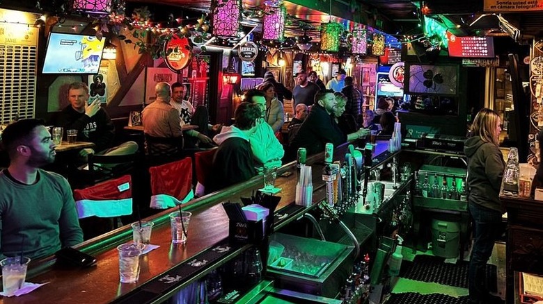 An angle view of the interior of a narrow, full, dimly-lit bar that's decorated with string lights and liquor/beer signs