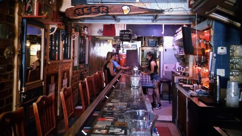 The interior of a narrow bar with cluttered walls and wood barstools as seen from the side;  it's empty except for a female bartender helping a man and a woman at the far end