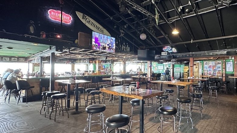 The interior of a large indoor-outdoor bar during the day with several empty tables with backless barstools and a square-shaped bar in the background; sparsely decorated except for posters tacked to the walls and a few beer signs
