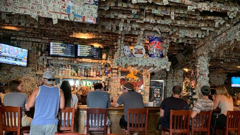 The interior of a bar with seven wood barstools filled with patrons as viewed from the back; walls, ceiling, and columns are covered in dollar bills