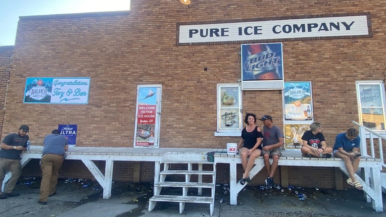 The exterior side of a large brick building with a white sign that says "Pure Ice Company" in black with a white wood loading dock with two men at the left side leaning on it and a family of four on the right side sitting on it
