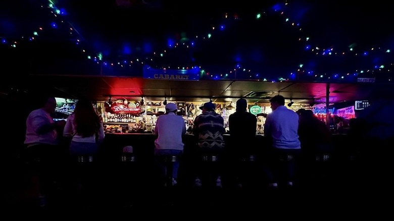 Seven bar patrons, as seen from the back, sitting on backless barstools face a lit, cluttered barback in a very dark bar decorated with multicolored string lights