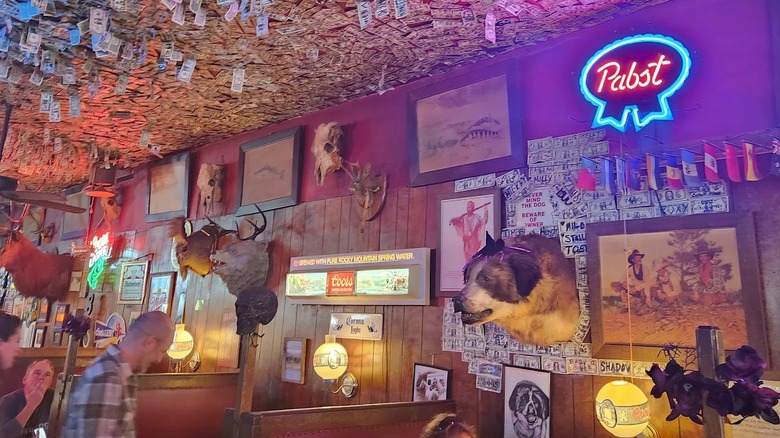The interior of a bar with taxidermy, beer signs, and random art as decoration on wood-paneled walls and dollar bills covering ceiling