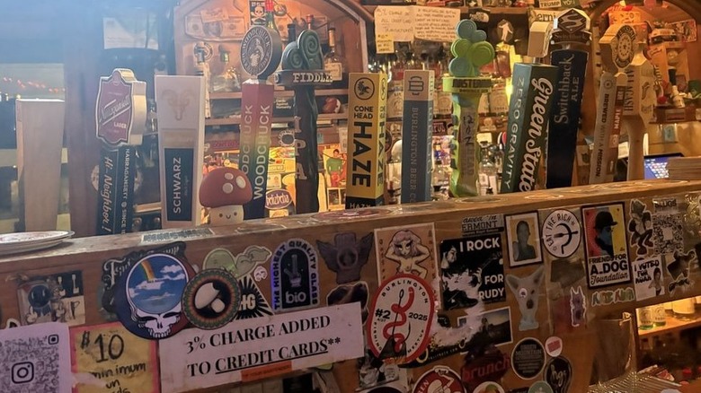 A line of 12 tap handles inside a bar behind a wood beam that is covered in various stickers; the back bar behind is filled with clutter