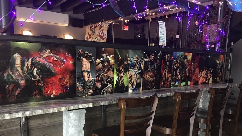 The interior of a bar showing a long, narrow, rough stone-topped table with a divider featuring seven scenes from GWAR concerts; purple string lights hang from the rafters
