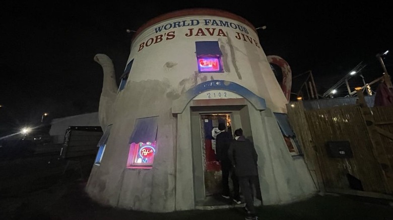 An evening shot of two people entering a concrete building that is shaped like a white teapot with a red lid; "World Famous Bob's Java Joint" is written on top and the are neon signs in the windows