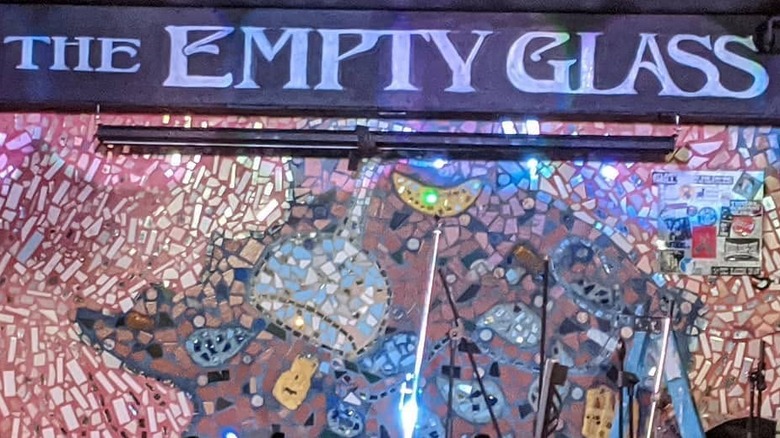 A bar stage with a multicolored modern art mosaic as the backdrop; "The Empty Glass" is written on a black background above it.