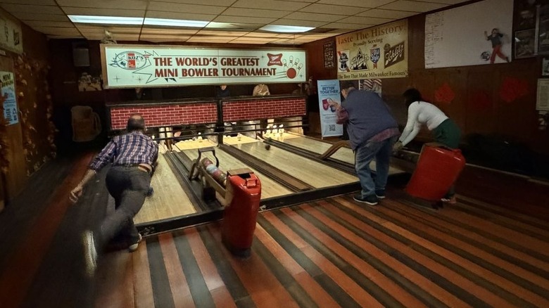 The bowlers bowl on three of four old-school duckpin bowling lanes; a sign that says "The World's Greatest Mini Bowler Tournament" is displayed at the far end of the lanes