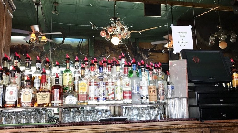 A wood back bar with a POS system on the right and glasses set in the forefront with two shelves of booze in the back; bar mirror has a prominent bullet hole in the center