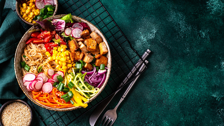 Vegan Buddha bowl with tofu, corn, peppers, onions, radishes, carrots, and quinoa on a green tablecloth with a fork and knife