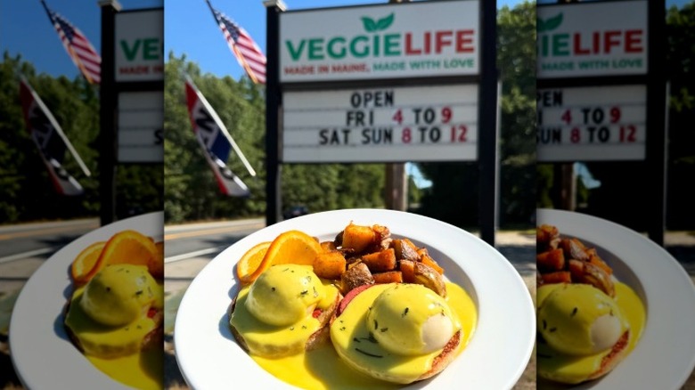A plate of vegan eggs benedict and potatoes held in front of signage for Veggie Life restaurant in Wells, Maine