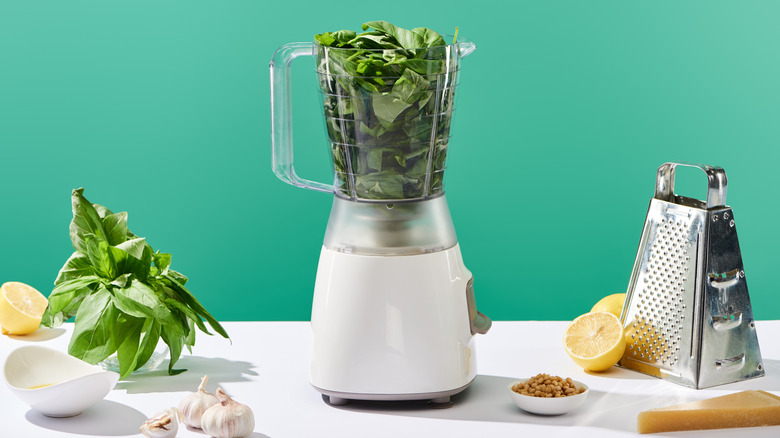 A blender full of fresh basil on a table with parmesan, lemon, and garlic
