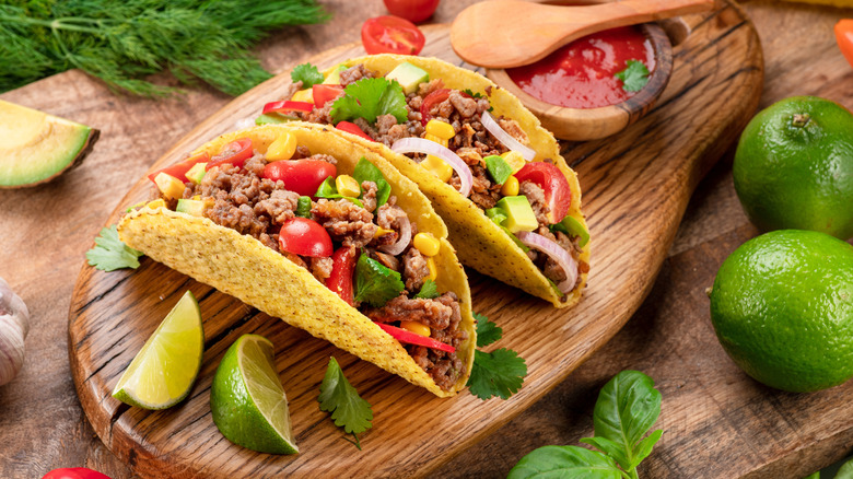 Two tacos with peppers in them rest on a wooden cutting board surrounded  by limes and a wooden dish of chili sauce