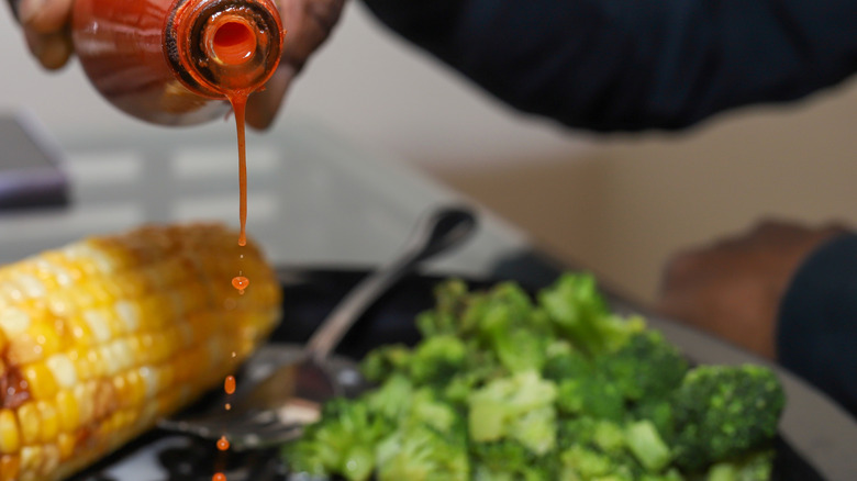 A person pouring hot sauce onto their plate of corn and broccoli