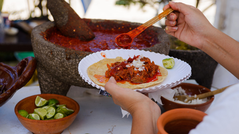A person scoops red sauce from a stone bowl onto food on a paper plate