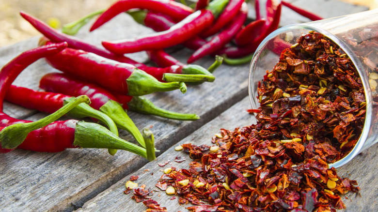 Red chili peppers sit beside a spilled glass of red chili pepper flakes on a wooden surface