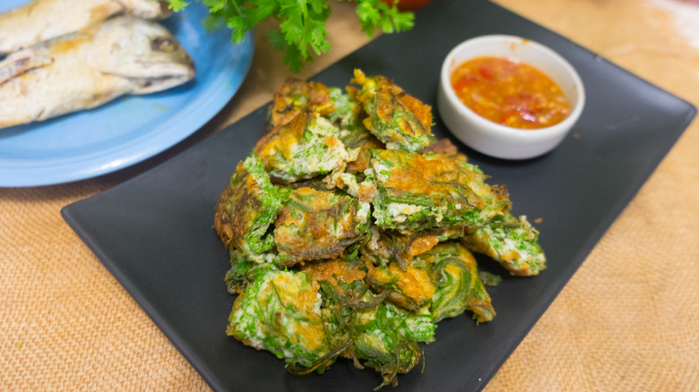 A spicy Thai food dish rests on a black rectangular plate with a dish of dipping sauce to the right