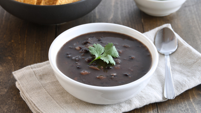 Black bean soup in a white bowl and a spoon on top of a cloth napkin and wood surface