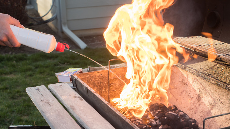 Pouring lighter fluid onto charcoal in a grill with rising flames from the briquettes.