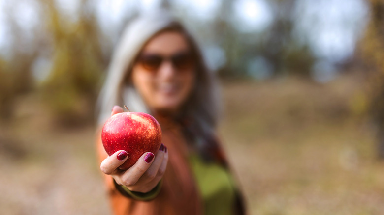 Woman holding a red apple in front of her with blurred trees behind her