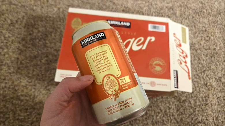 A hand holding a Kirkland Signature Lager can, showing the back text about the origin of the beer, with a box of beer in the background