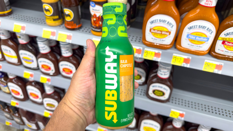 A hand holds a bottle of Subway Baja Chipotle sauce in front of grocery store condiment shelves.