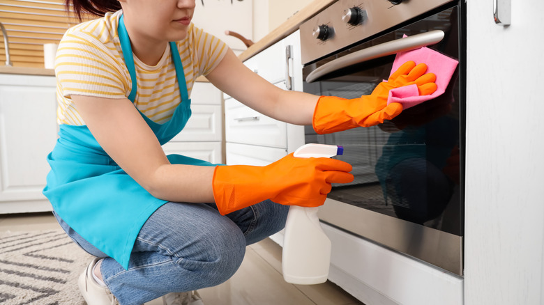 Person cleaning a glass oven door with spray bottle and cloth