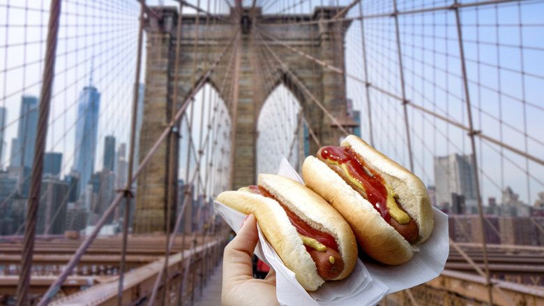 Hand holding two hot dogs in front of the Brooklyn Bridge.