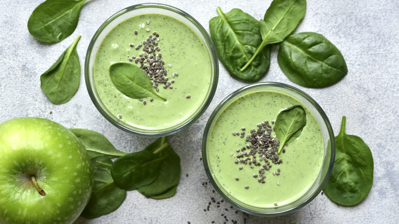 Two glasses of green smoothie surrounded by spinach leaves and an apple