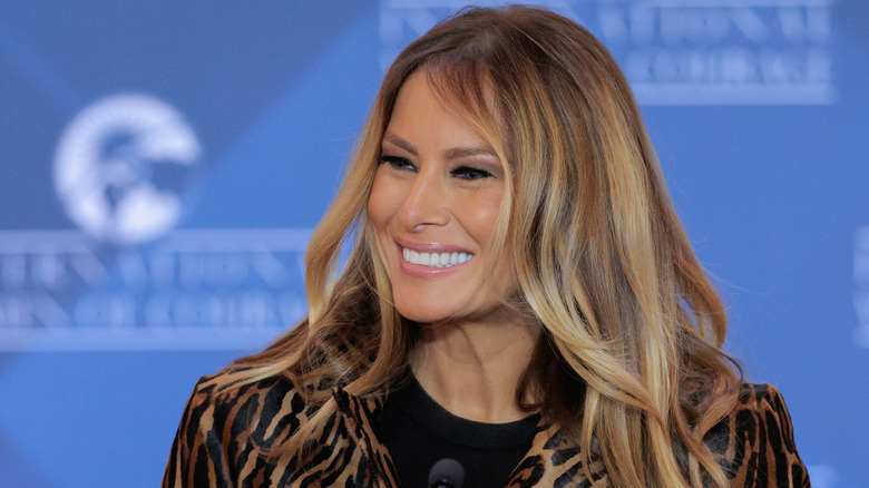 Melania Trump looking to the side and smiling in front of a blue background