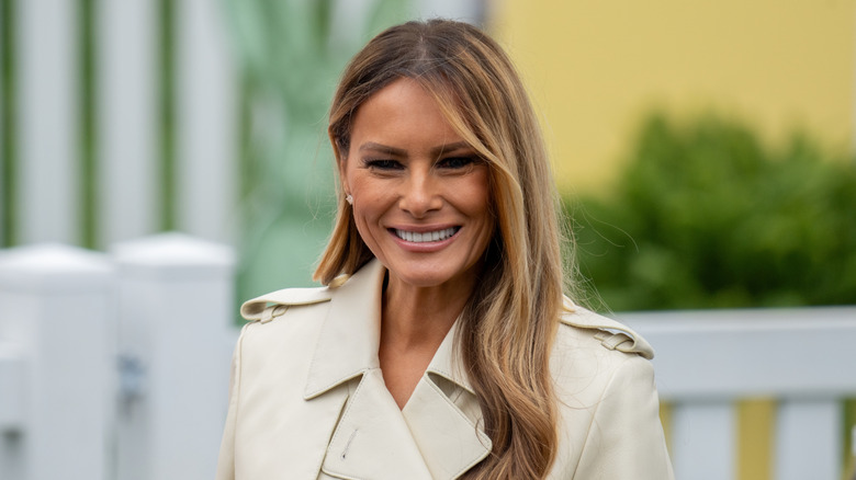Melania Trump smiles at the camera wearing a tan coat