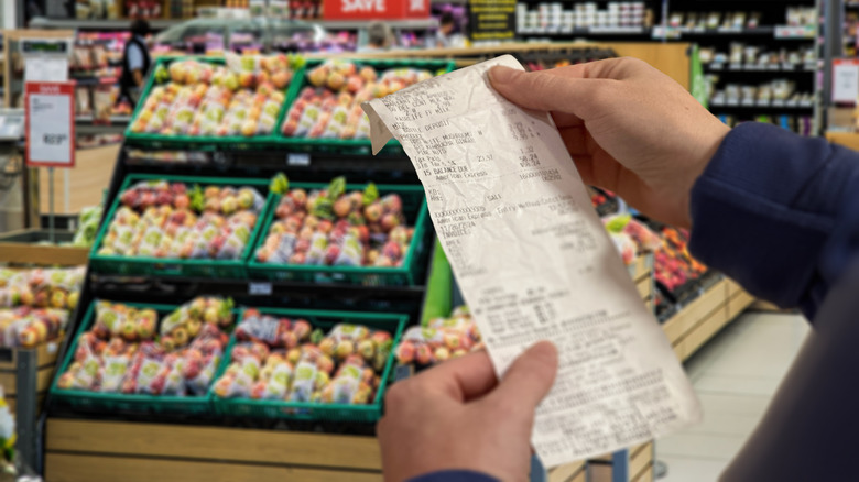 Hands holding up a grocery store receipt in the foreground with various grocery products in the background