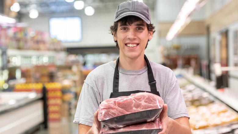 A young man at a grocery store holding packages of meat and smiling.