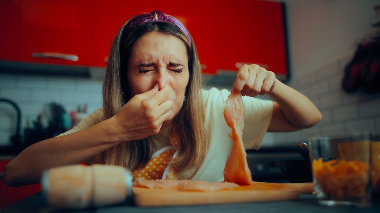 Woman holding her nose while holding a piece of chicken that's apparently spoiled.