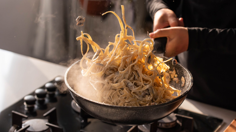 A cook tossing a pan of steaming fettuccine pasta mid air with creamy mushroom sauce on a stovetop.