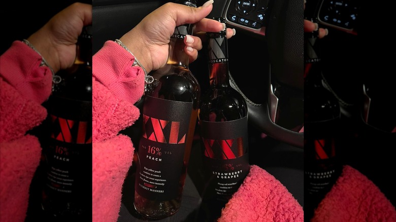 A woman wearing a pink fuzzy jacket holds two bottles of XXL wine in her car