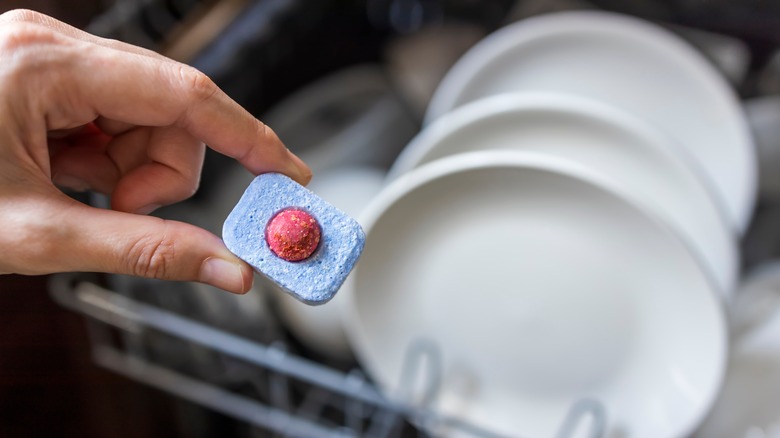 A closeup of a hand holding a dishwasher detergent tablet in front of an open dishwasher with plates