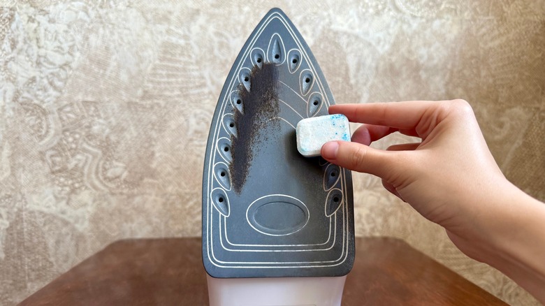 A hand using a dishwasher tablet to remove stuck-on burned residue from the underside of an iron