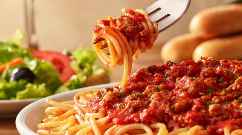A fork lifting spaghetti with meat sauce form Olive Garden with breadsticks and salad in background