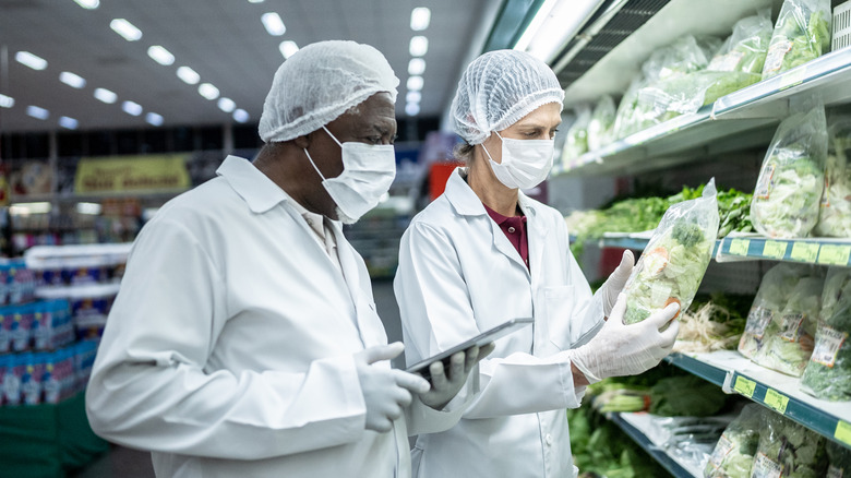 Two health inspectors looking at bags of greens in a grocery store with protective gear on.
