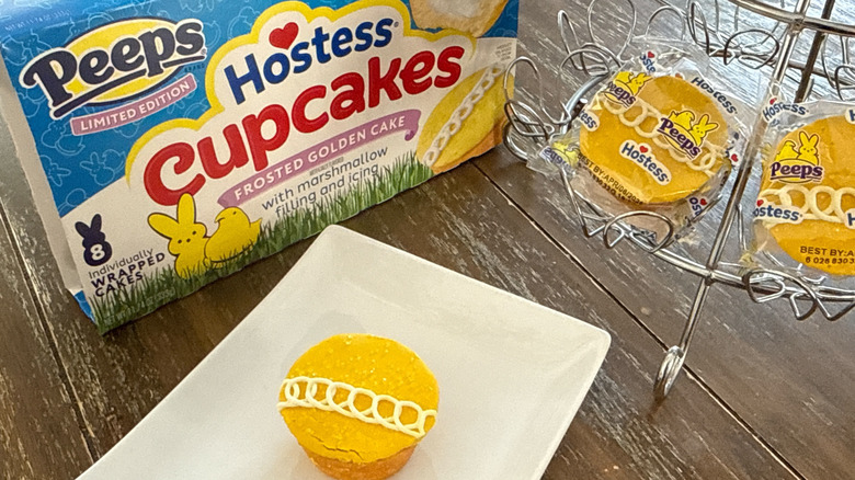Hostess Peeps cupcake on white plate with wrapped ones in a silver cupcake stand with product box behind