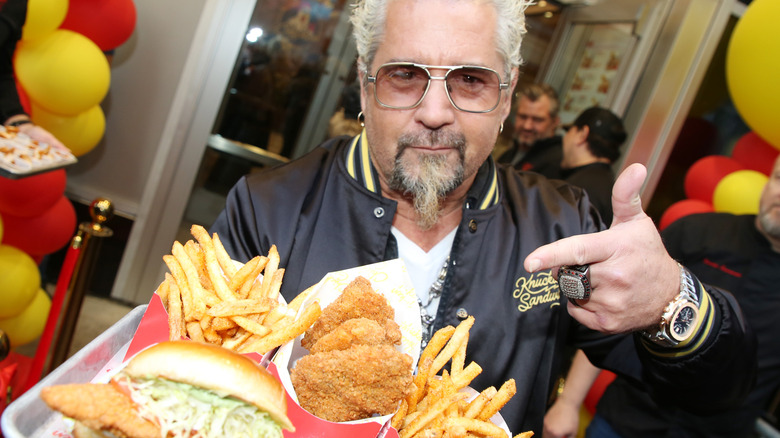 Guy Fieri wearing sunglasses pointing to a tray of food.