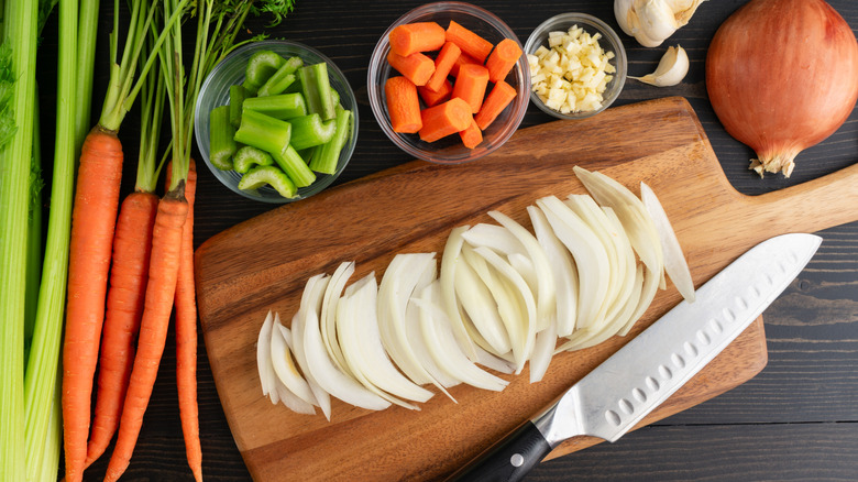Sliced onions on a cutting board with whole and chopped celery, carrots, and garlic plus a knife