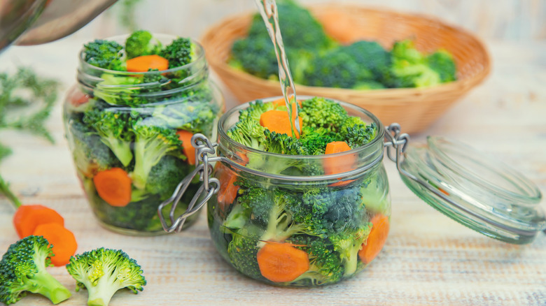 White vinegar being poured into jars of fresh broccoli and carrots