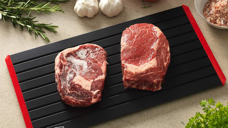 Two slices of meat placed above a black and red defrosting tray, surrounded by other ingredients on a kitchen table
