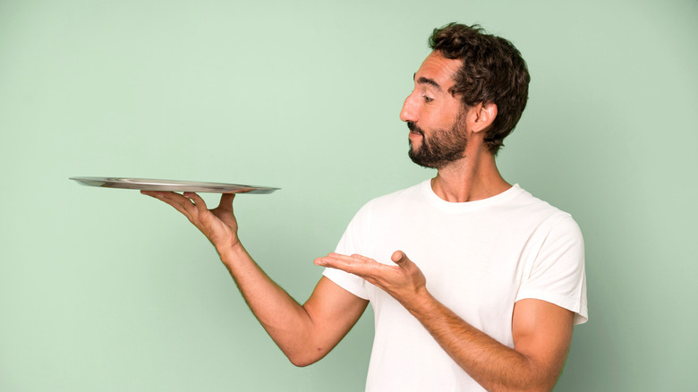 A bearded man in white shirt holding an empty steel try with one hand.