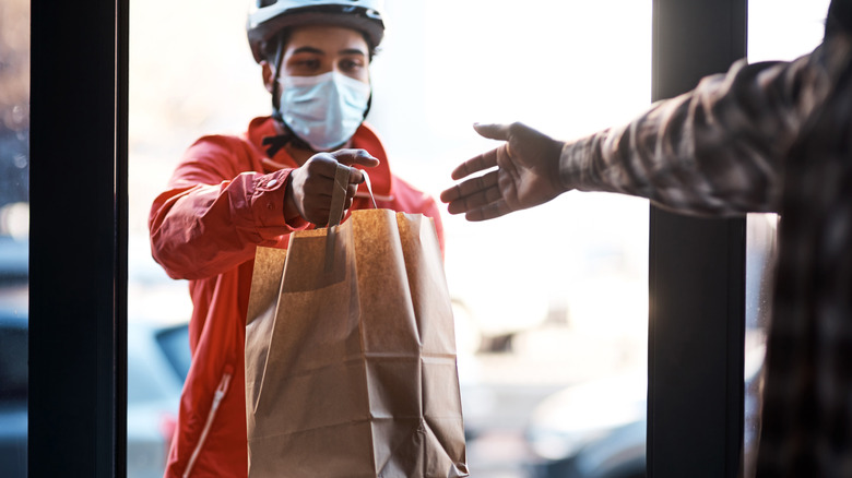 A delivery driver with a mask on hands a bag of food to a customer off screen.