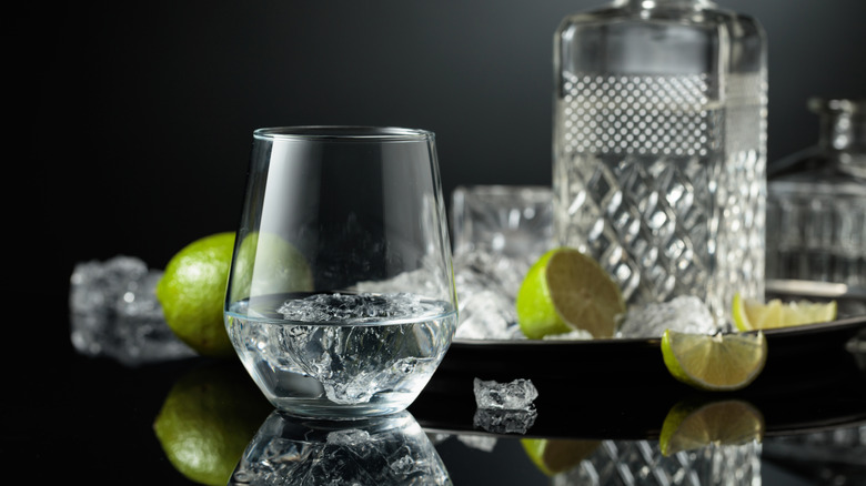 glass of vodka with large ice cube, ice and limes cut and whole