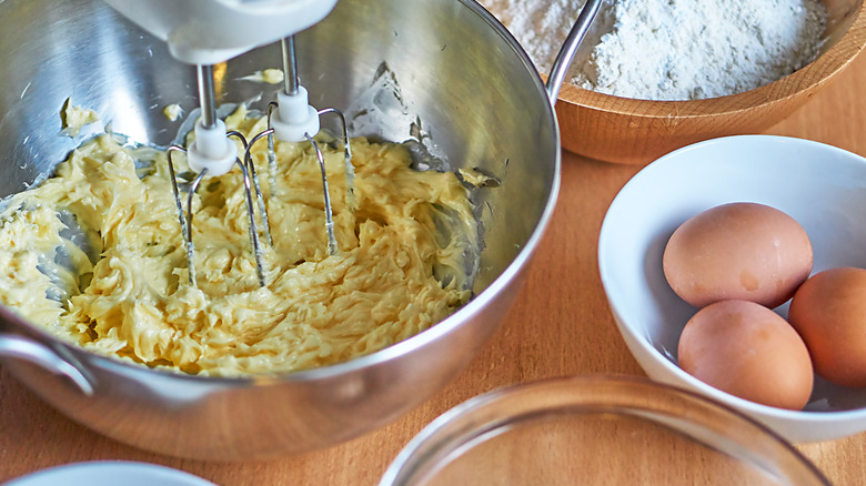 Yellow cake batter being beaten in a metal bowl with an electric mixer, with a small bowl with three brown eggs next to it